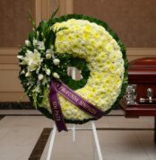 Corona fúnebre Bethesda con flores blancas y amarillas, cinta morada con mensaje 'En Profunda Simpatía', junto a un ataúd en