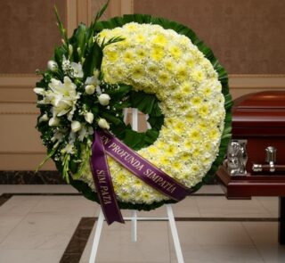 Corona fúnebre Bethesda con flores blancas y amarillas, cinta morada con mensaje 'En Profunda Simpatía', junto a un ataúd en