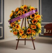Corona fúnebre de girasoles vibrantes con cinta morada que dice 'Nuestro Más Sentido Pésame', exhibida en una sala de velació