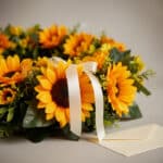 Fotografía macro premium de una corona fúnebre con girasoles vibrantes, follaje verde y una cinta satinada blanca. Los pétalo