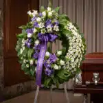 Corona fúnebre 'Homenaje a Daniel' con flores blancas y moradas, cinta morada con mensaje, en sala de velación elegante junto