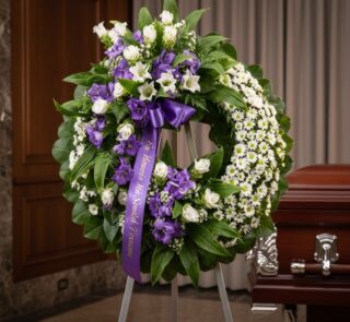 Corona fúnebre 'Homenaje a Daniel' con flores blancas y moradas, cinta morada con mensaje, en sala de velación elegante junto