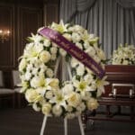 Corona fúnebre Homenaje Gadriel con flores blancas, lirios y rosas, sobre un caballete blanco. Cinta morada con el mensaje 'N