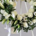 Corona fúnebre 'Homenaje Sincero' con lirios, rosas y crisantemos blancos, follaje verde, rocío sutil, cinta satinada y sobre