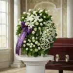 Corona fúnebre Legado Jacob con rosas blancas, lirios y margaritas, cinta morada con 'Deepest Sympathy', junto a un ataúd en