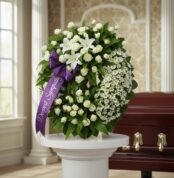 Corona fúnebre Legado Jacob con rosas blancas, lirios y margaritas, cinta morada con 'Deepest Sympathy', junto a un ataúd en