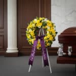 Corona fúnebre de flores amarillas y blancas con cinta morada "En Profunda Simpatía" junto a un ataúd en sala de velación ele