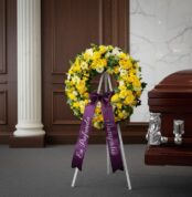 Corona fúnebre de flores amarillas y blancas con cinta morada "En Profunda Simpatía" junto a un ataúd en sala de velación ele