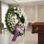 Corona fúnebre Legado de Paz con flores blancas, liliums, margaritas y anturios, cinta morada 'En Paz Descanse' en sala de ve