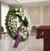 Corona fúnebre Legado de Paz con flores blancas, liliums, margaritas y anturios, cinta morada 'En Paz Descanse' en sala de ve