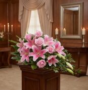 Corona fúnebre Lino con rosas rosadas y lirios, exhibida elegantemente en una sala de velación con iluminación cálida y fondo