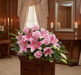 Corona fúnebre Lino con rosas rosadas y lirios, exhibida elegantemente en una sala de velación con iluminación cálida y fondo