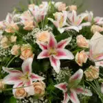 Primer plano de una corona fúnebre 'Luz Eterna' con lirios blancos y rosas melocotón, rocío sutil, cinta satinada y follaje v