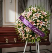 Corona fúnebre Luz Eterna con lirios blancos y rosas melocotón, cinta morada 'En Memoria Amorosa', junto a un ataúd de madera
