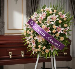 Corona fúnebre Luz Eterna con lirios blancos y rosas melocotón, cinta morada 'En Memoria Amorosa', junto a un ataúd de madera