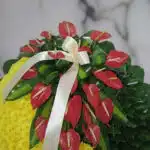 Fotografía macro premium de la Corona Fúnebre a Tu Memoria, mostrando anthuriums rojos, follaje verde y una cinta satinada co