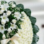 Primer plano de una corona fúnebre con rosas blancas, lirios y crisantemos, con una cinta satinada color crema y follaje verd