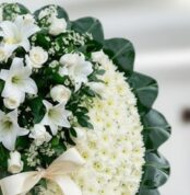 Primer plano de una corona fúnebre con rosas blancas, lirios y crisantemos, con una cinta satinada color crema y follaje verd