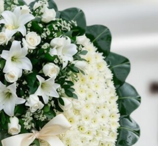 Primer plano de una corona fúnebre con rosas blancas, lirios y crisantemos, con una cinta satinada color crema y follaje verd