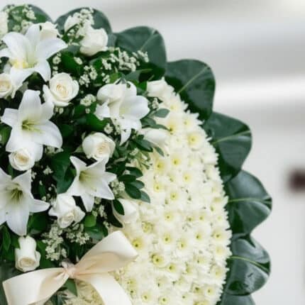 Primer plano de una corona fúnebre con rosas blancas, lirios y crisantemos, con una cinta satinada color crema y follaje verd