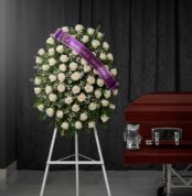 Corona fúnebre de rosas blancas y follaje verde, con una cinta morada que dice 'En memoria cariñosa', junto a un ataúd de mad