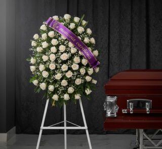 Corona fúnebre de rosas blancas y follaje verde, con una cinta morada que dice 'En memoria cariñosa', junto a un ataúd de mad
