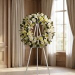 Corona fúnebre "Solemne Homenaje Joshua" con lirios blancos, rosas y crisantemos, sobre un caballete en una sala de velación