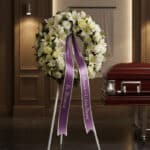 Corona fúnebre de flores blancas y verdes con cinta morada, "En Memoria" y "Profundo Pésame", junto a un ataúd en sala de vel
