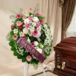 Imponente corona fúnebre "Homenaje a Mathias" con lirios blancos y rosados, gerberas y follaje verde, junto a un ataúd en sal