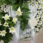 Detalle macro de corona funeraria con lirios blancos, margaritas frescas y follaje verde, con cinta satinada y rocío sutil, p