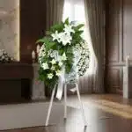 Corona de homenaje "El Vuelo de Uriel" con lirios blancos, margaritas y follaje verde, sobre un trípode blanco en una sala de