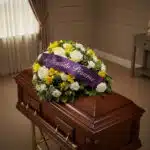 Cubre caja fúnebre Baltasar con rosas blancas, fresias amarillas y follaje verde, adornado con una cinta morada que dice 'Con