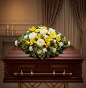 Cubre Caja Fúnebre Baltasar con rosas blancas, freesias amarillas y margaritas, sobre un ataúd de madera oscura en un entorno