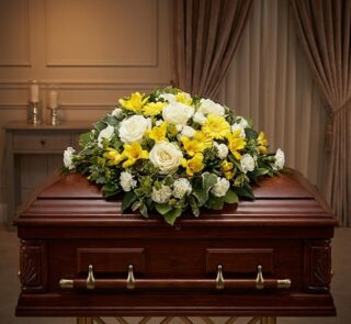 Cubre Caja Fúnebre Baltasar con rosas blancas, freesias amarillas y margaritas, sobre un ataúd de madera oscura en un entorno