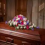 Cubre caja fúnebre "El Legado de Francisco" con flores variadas en tonos rosas, morados y blancos, sobre un ataúd de madera o