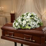 Cubre caja fúnebre Epifania con flores blancas y verdes sobre un ataúd de caoba brillante en una sala de velación de lujo.