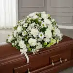 Cubre caja fúnebre Epifania con flores blancas y cinta de condolencia, vista desde un ángulo diferente y composición renovada