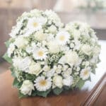 Primer plano de un cubre caja fúnebre con rosas blancas, lirios y claveles, con iluminación celestial y etérea, transmitiendo