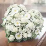 Primer plano de un cubre caja fúnebre con rosas blancas, lirios y claveles, con iluminación celestial y etérea, transmitiendo