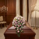 Cubre caja fúnebre con rosas rosadas y follaje verde, sobre un ataúd de madera oscura en una sala de velación premium con luz