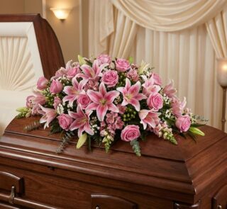 Cubre caja fúnebre Ifigenia con rosas y lirios rosados, eucalipto y follaje verde, elegantemente dispuesto sobre un ataúd de