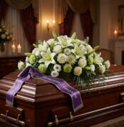 Cubre caja fúnebre "Legado Eterno de Tomás" con lirios, rosas blancas y cinta morada en sala de velación premium, luz difusa.