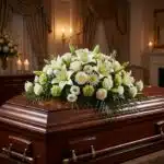 Cubre caja fúnebre 'Legado Eterno de Tomás' con lirios blancos, rosas y follaje verde sobre un ataúd de madera oscura en un a