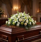 Cubre caja fúnebre 'Legado Eterno de Tomás' con lirios blancos, rosas y follaje verde sobre un ataúd de madera oscura en un a