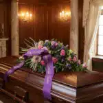 Cubre caja fúnebre Legado Sereno de Ivon con flores rosas y moradas, y una cinta morada con el mensaje 'En Memoria' sobre un