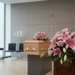 Cubre caja fúnebre Lino con rosas y lirios rosados en primer plano, en una sala de velación moderna con ataúd de madera desen