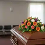 Cubre caja fúnebre 'Paz Eterna' con flores variadas en tonos naranja, blanco y morado, sobre un ataúd de madera en una sala d