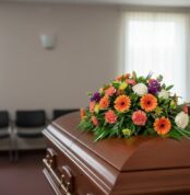Cubre caja fúnebre 'Paz Eterna' con flores variadas en tonos naranja, blanco y morado, sobre un ataúd de madera en una sala d