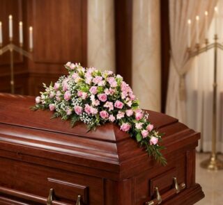 Cubre caja fúnebre elegante con rosas rosadas y follaje verde, delicadamente dispuesto sobre un ataúd de madera oscura en un