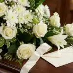 Primer plano de un cubre caja fúnebre con rosas blancas, lirios y crisantemos, con rocío sutil y una cinta satinada color cre